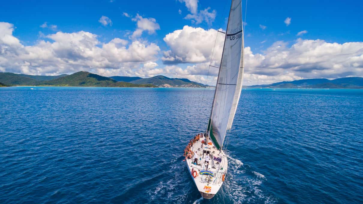 A white maxi yacht heeling under sail on deep blue water with a green Whitsundays island behind
