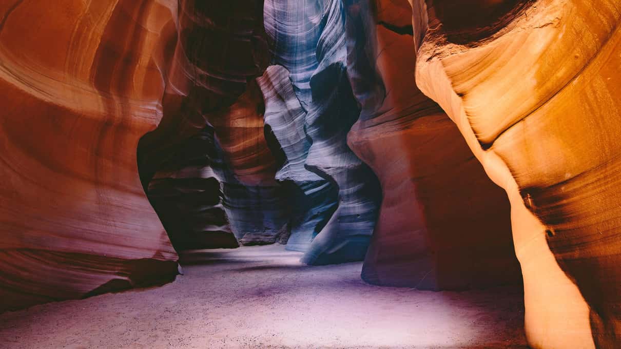 Sandy corridor of Upper Antelope Canyon in violet and orange light, opening toward a bright gap
