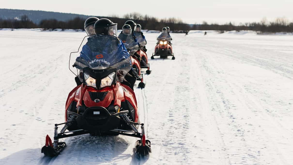 A long column of snowmobiles crossing a wide, snow-covered frozen lake on a Safartica safari near Rovaniemi
