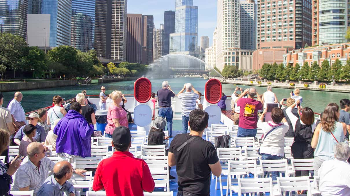 Passengers of all ages on the sunny open deck of a river boat with skyscrapers on both banks