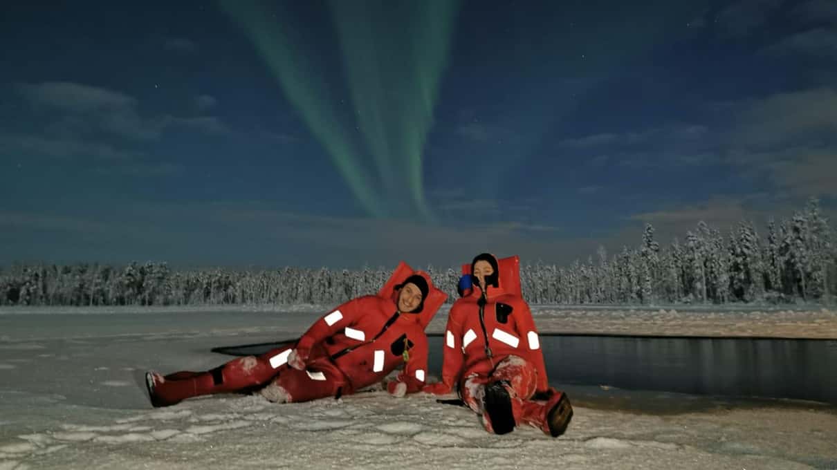 Two floaters in red suits lying on the ice under green northern lights at night