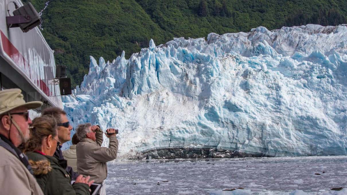 Passengers on a cruise deck raising binoculars toward the pale blue seracs of a tidewater glacier face