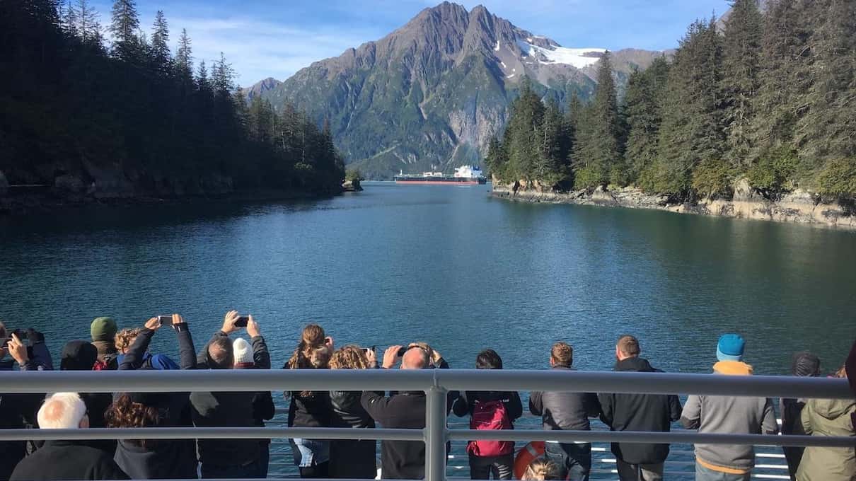 Passengers lining the stern rail of a tour boat as it runs a forested channel toward a bare grey peak