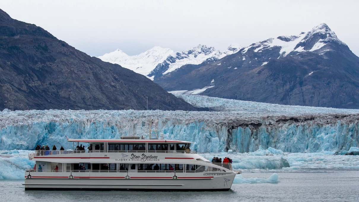 A white Stan Stephens tour boat passing a long blue ice cliff below snow-covered Chugach peaks