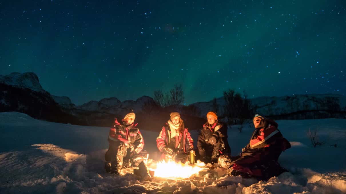 Cuatro personas sentadas alrededor de una hoguera sobre la nieve, de noche, con montañas y un leve resplandor de aurora en el cielo