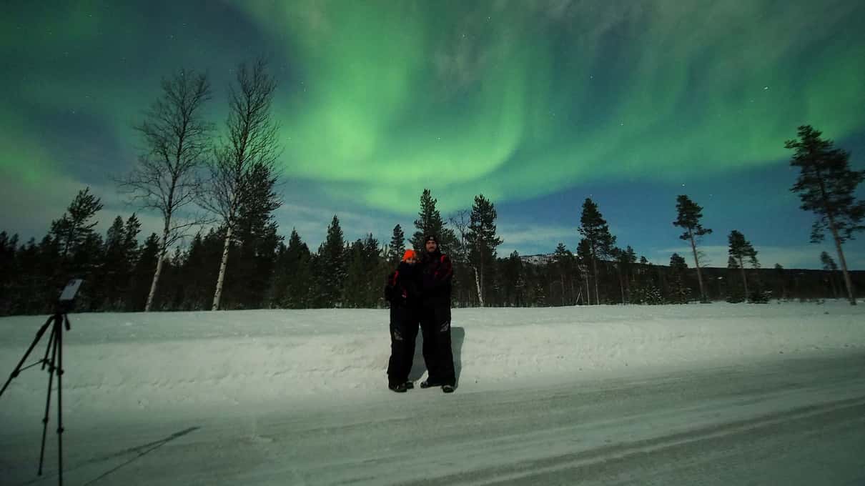 A green aurora arching over snowy birches as two guests stand beside a camera tripod