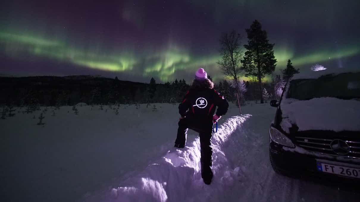 A guest with a headlamp walking along a snowy track beside a minibus under a purple and green aurora