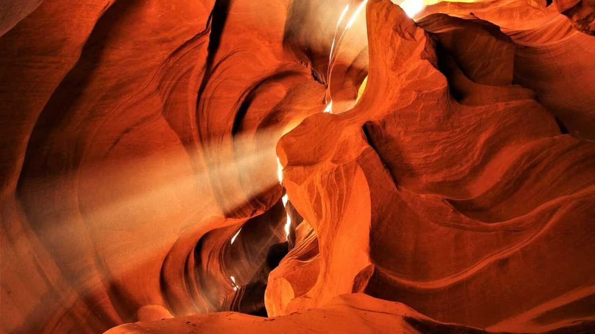 A single light beam full of falling sand dropping to the floor of Upper Antelope Canyon between dark curved walls
