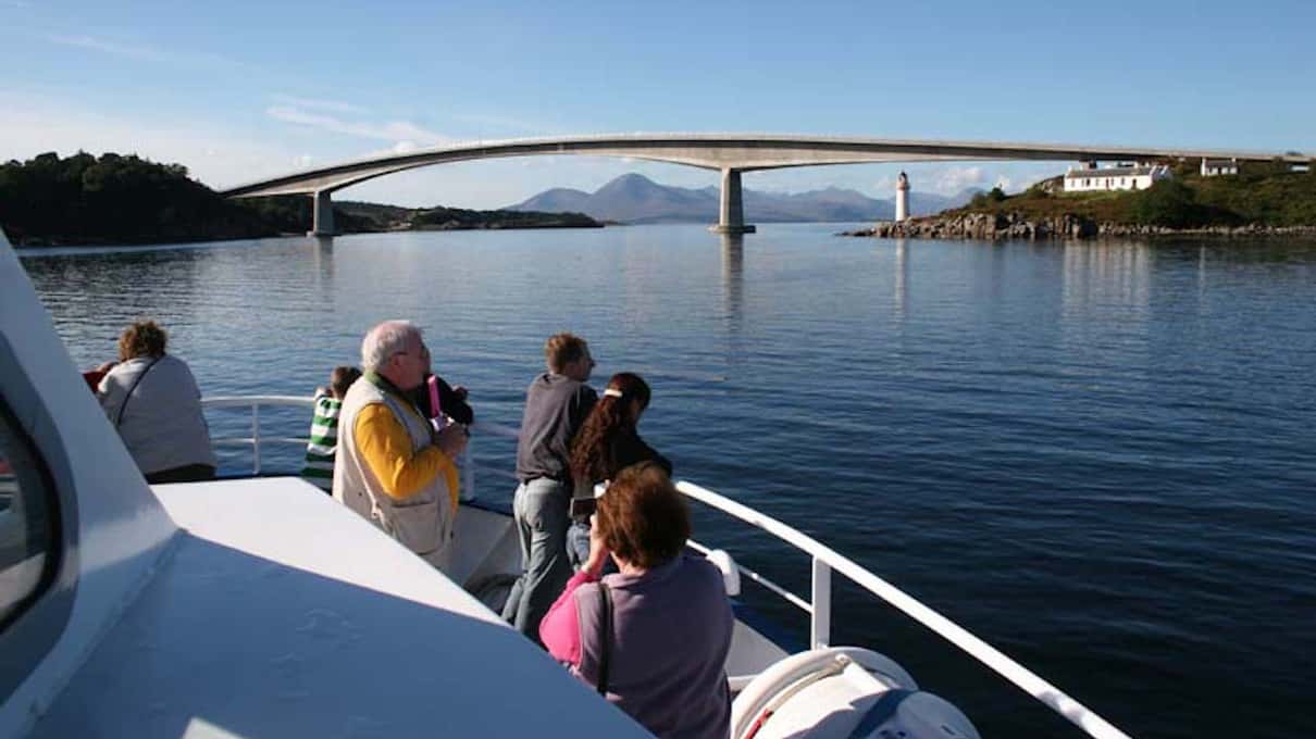 Passengers on the open deck of a boat looking toward the arch of the Skye Bridge and the white Kyleakin lighthouse