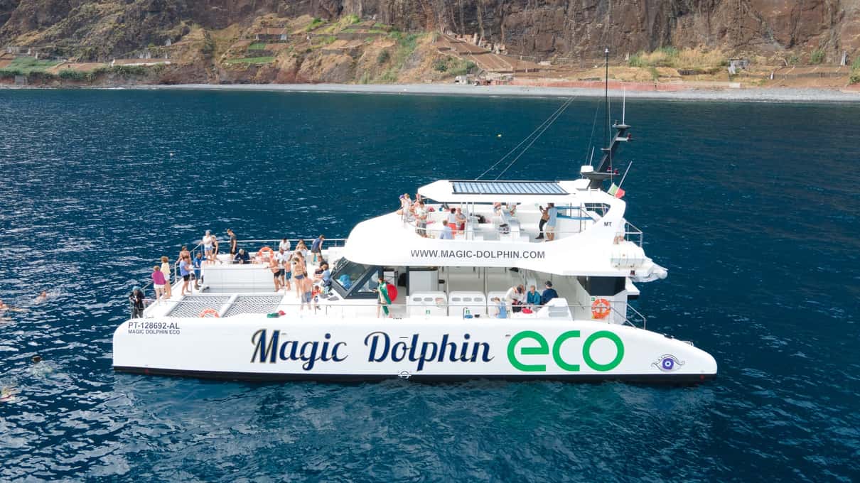 The white Magic Dolphin Eco catamaran full of guests on both decks below the dark cliffs of Cabo Girão