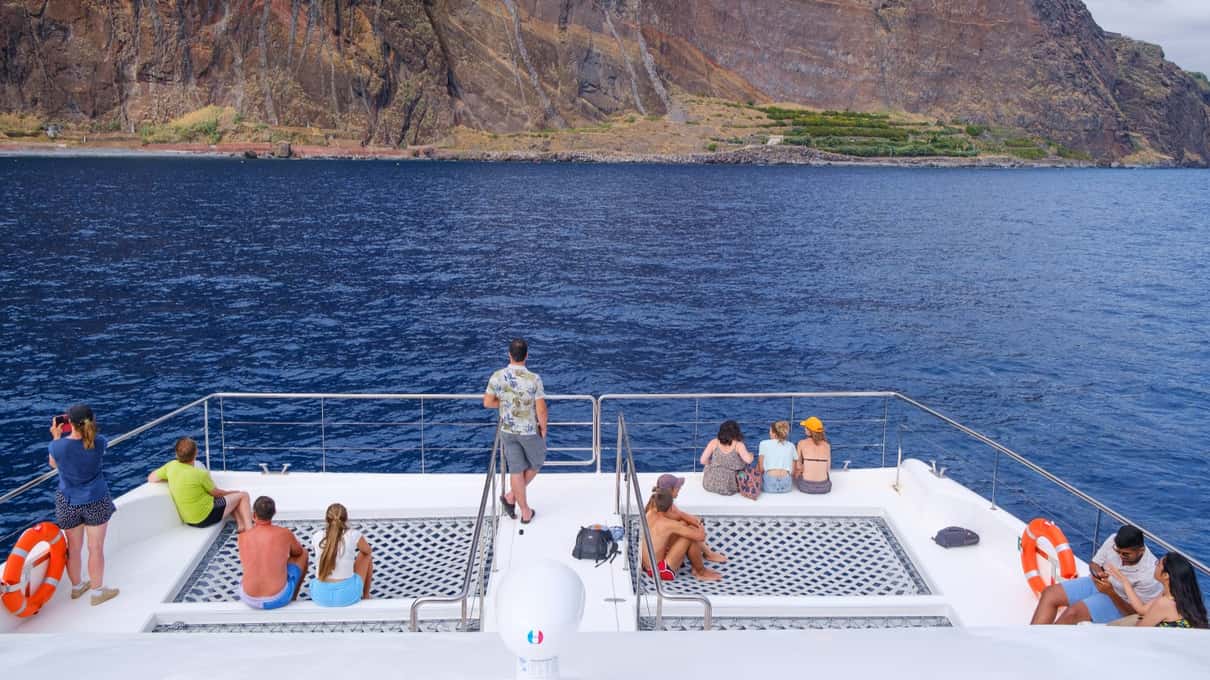 Guests sitting on the bow nets of a catamaran facing the dark sea cliffs of Cabo Girão