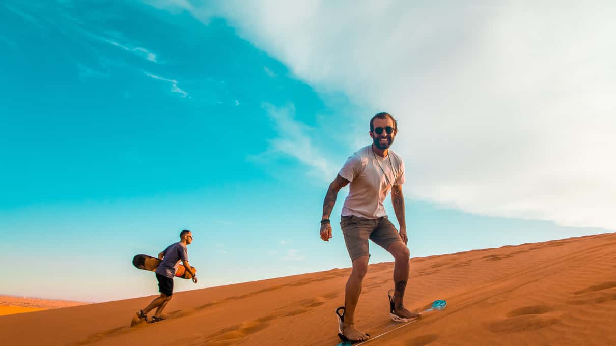 Reiziger glijdt op een sandboard een rode duin af onder een strakblauwe lucht, achter hem loopt een tweede met zijn board