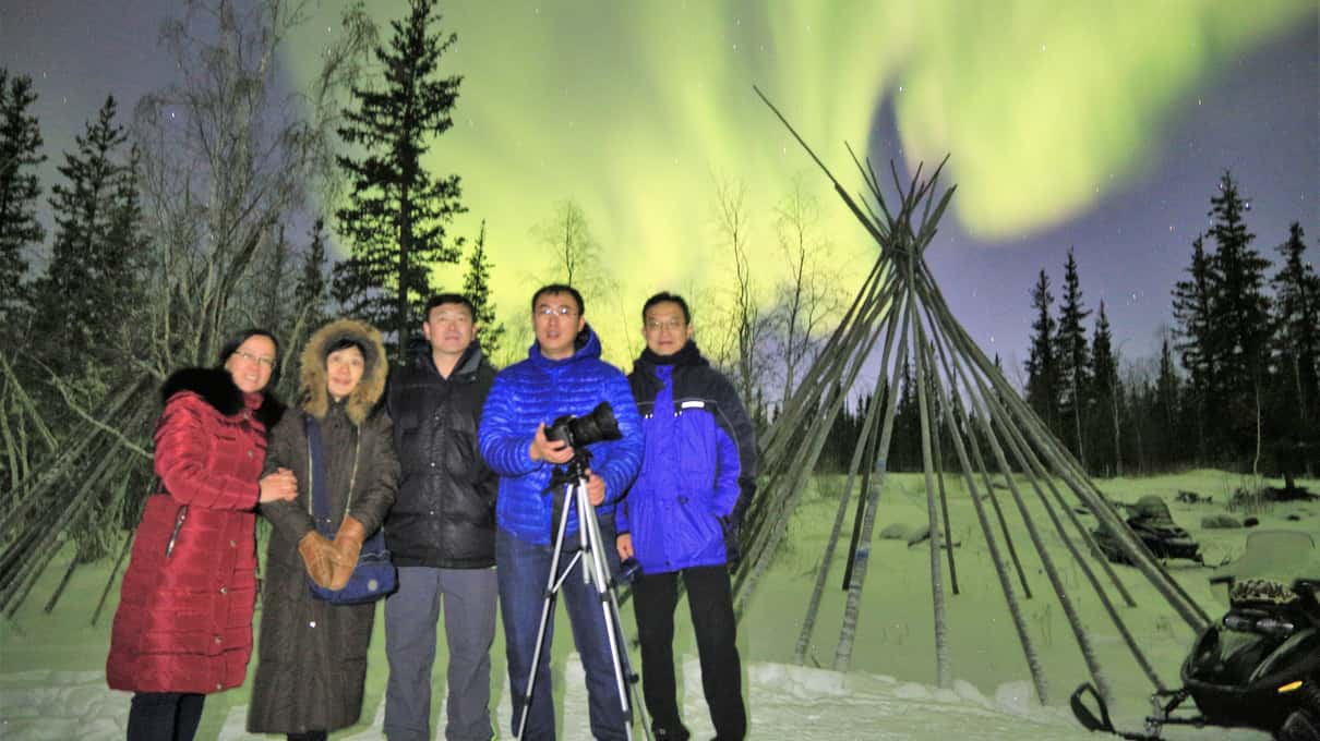 Five guests in winter parkas posing beside a wooden teepee frame under a green aurora near Yellowknife