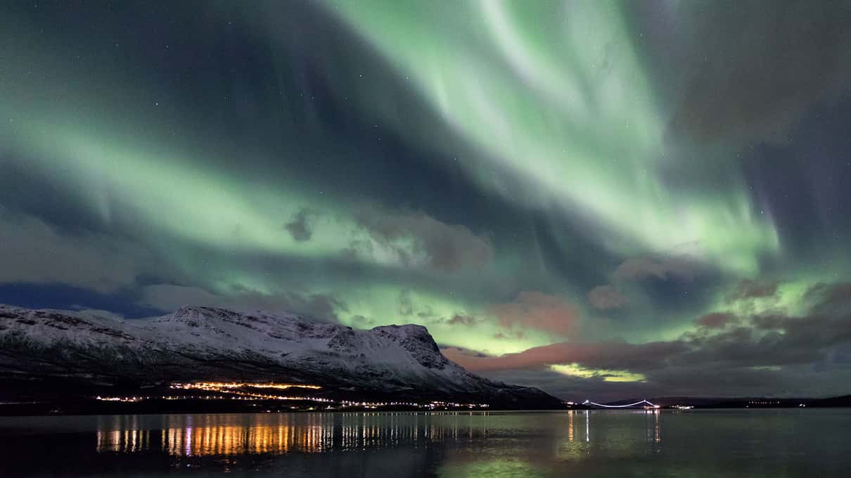 Aurora over Lake Torneträsk with village lights and snowy mountains on the far shore