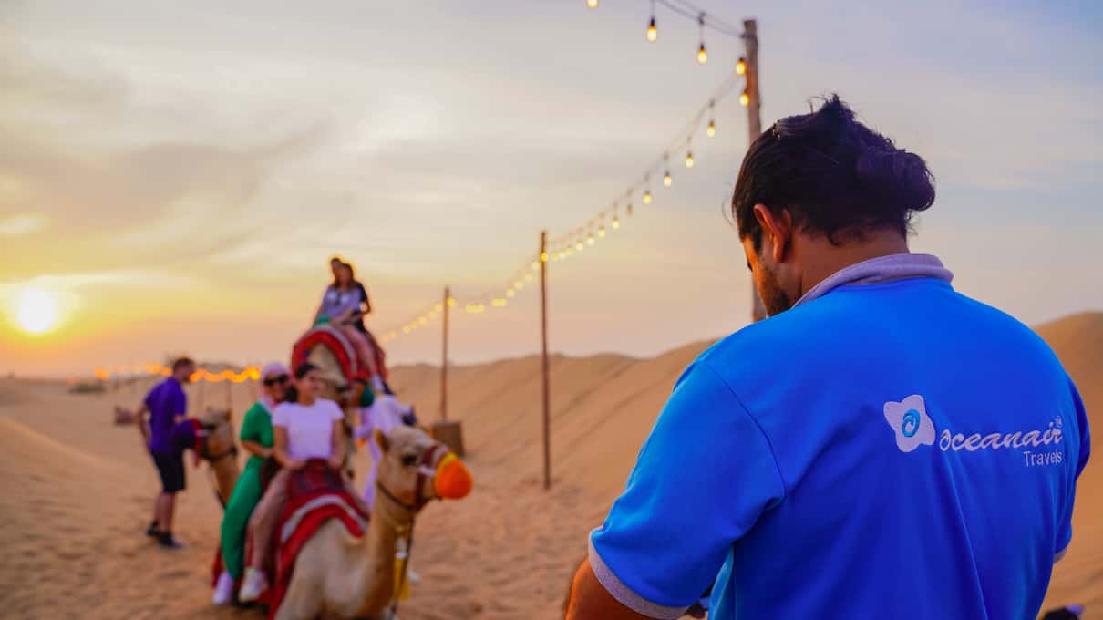 Viajeros montados en camello al atardecer en el campamento, bajo una guirnalda de luces, con un miembro del equipo de espaldas en primer plano