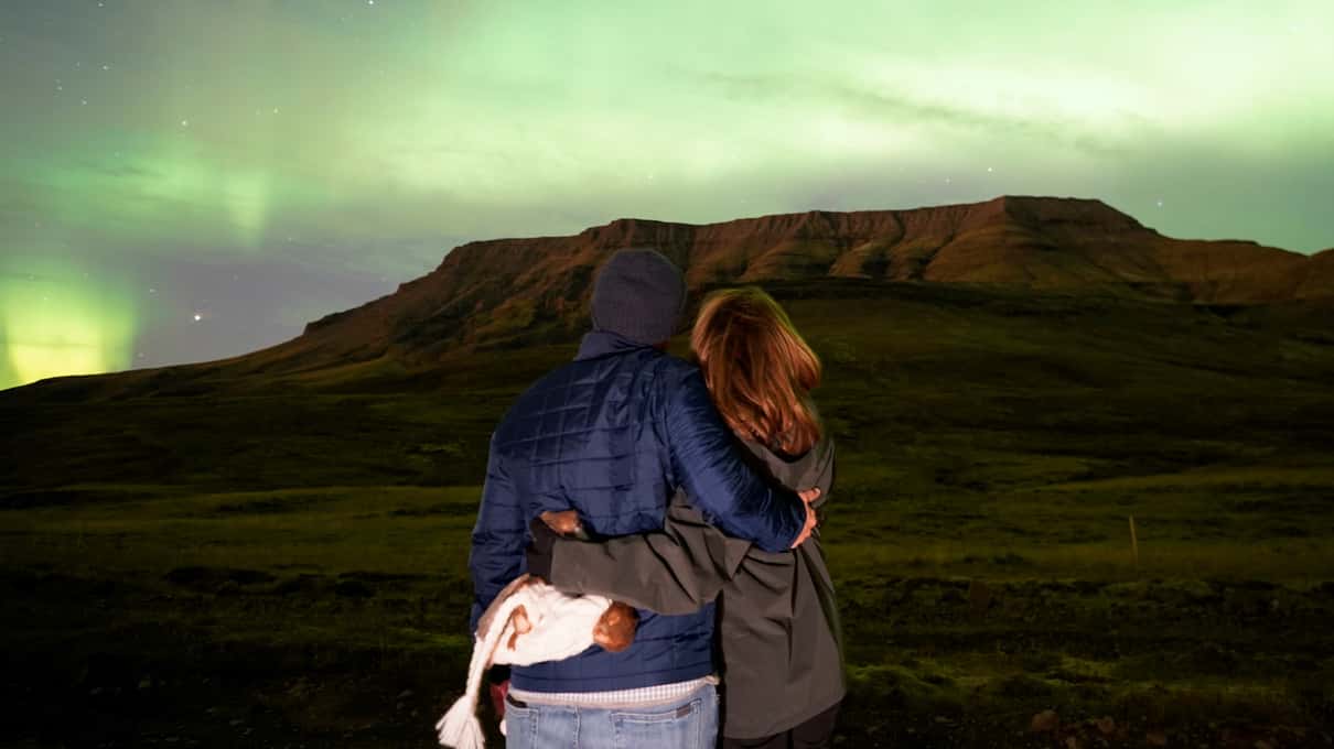 A couple hugging on snow-free grass under a green aurora above a flat-topped mountain in Iceland