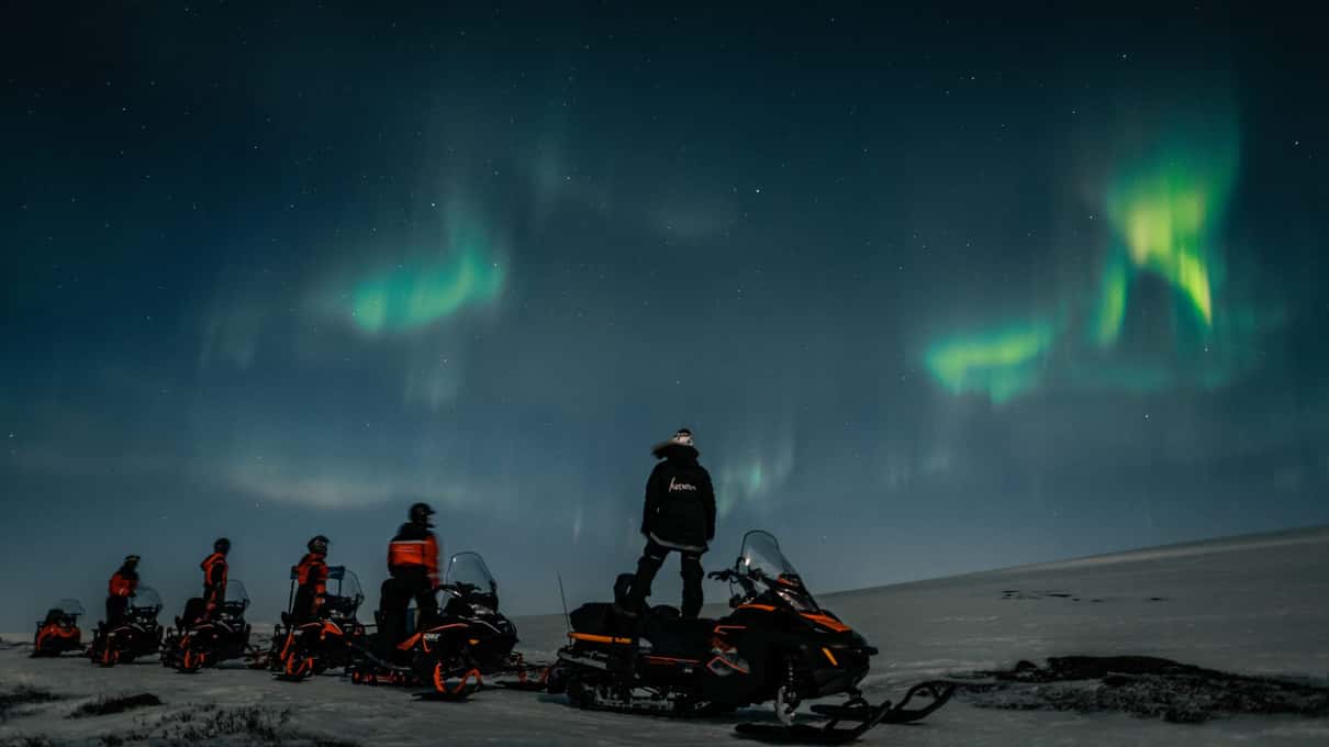 A row of snowmobiles on open snow with one rider standing under green aurora patches
