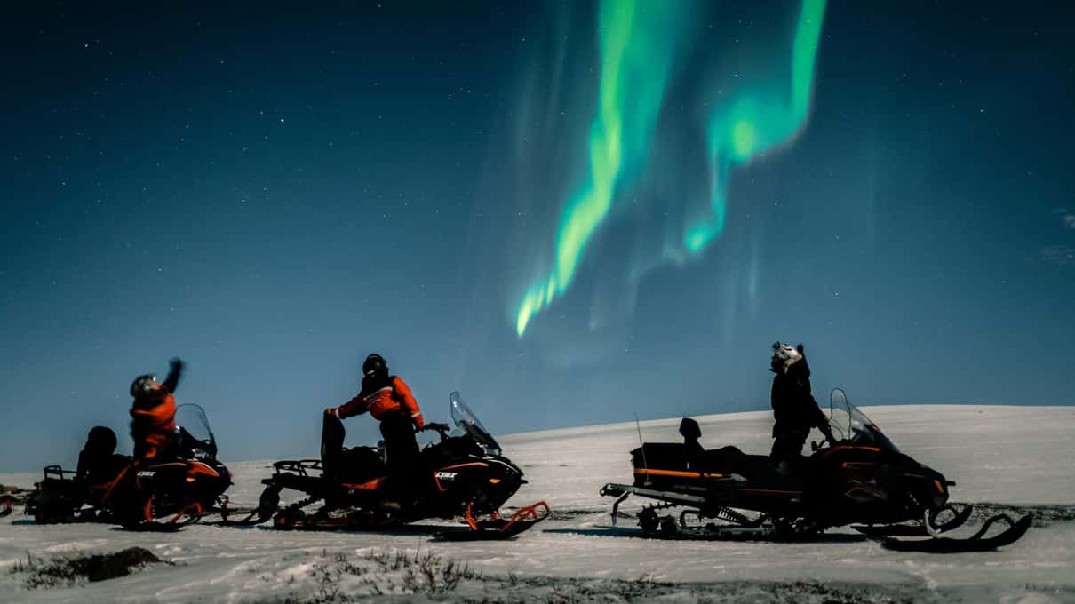 A green aurora arching over riders standing beside their snowmobiles at night on an Æventyr ride above Alta