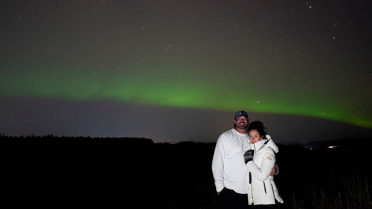 Two guests in white winter coats and hats standing under a low green aurora band at night