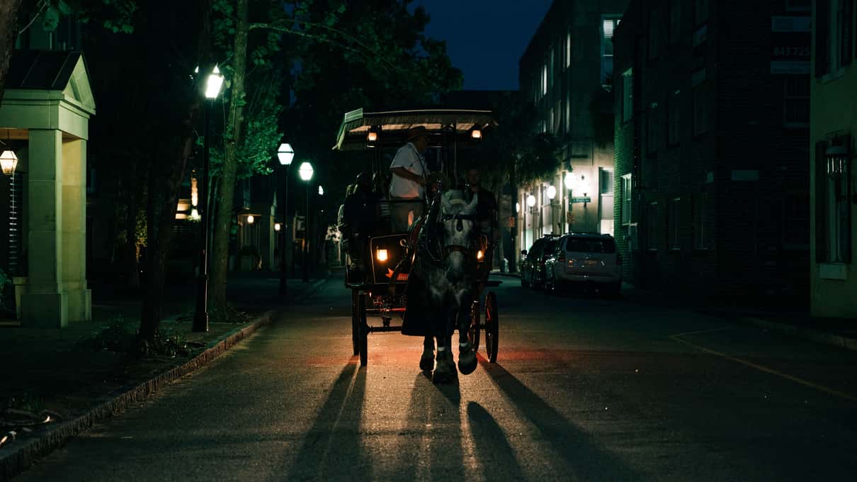 A horse and carriage seen in silhouette under glowing street lamps on a dark Charleston street