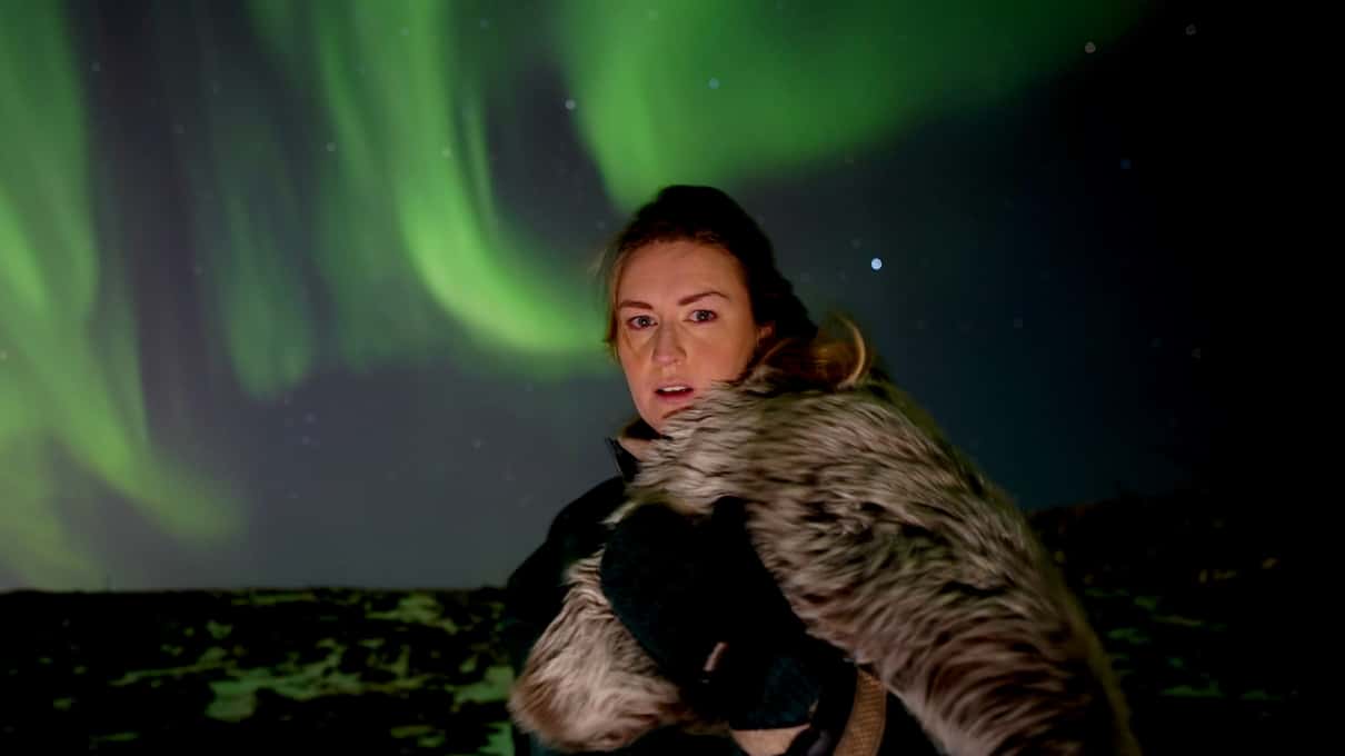 A guest wrapped in a Viking fur cape looking into the camera under a bright green aurora at night