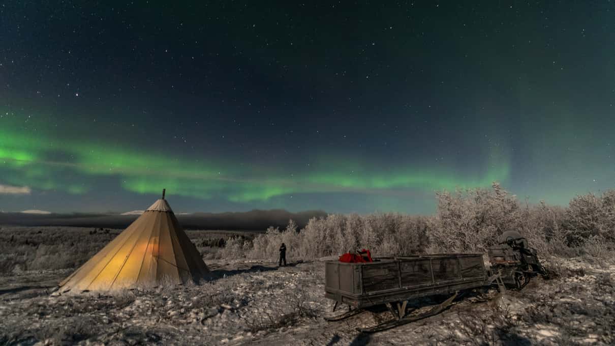 A lit canvas lávvu and a wooden sleigh on frosty ground under a faint green aurora