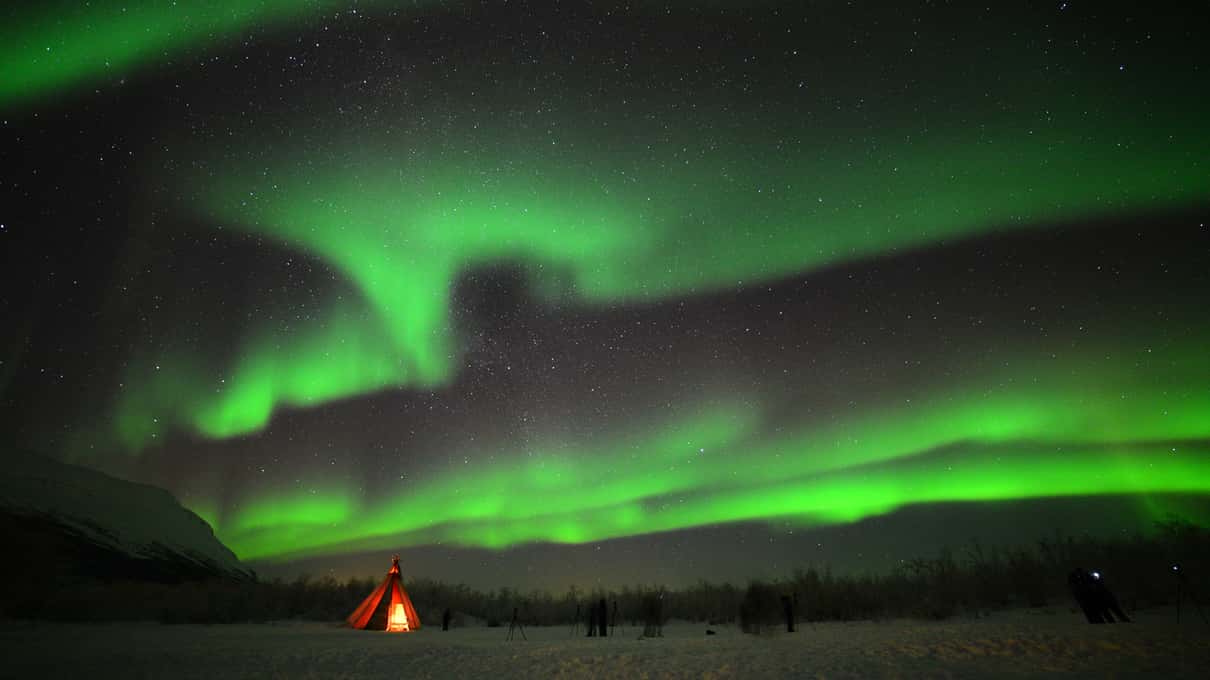 Green aurora bands over a dark snowy valley with a small lit tent far off on the snow