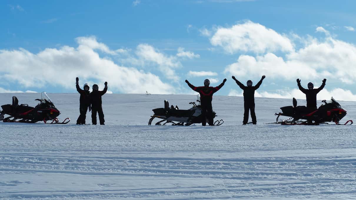 Riders with arms raised beside their snowmobiles under a wide sky on the plateau near Alta