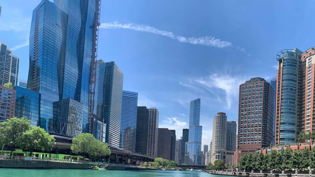 Glass towers lining the Chicago River on a sunny day with green trees along the bank