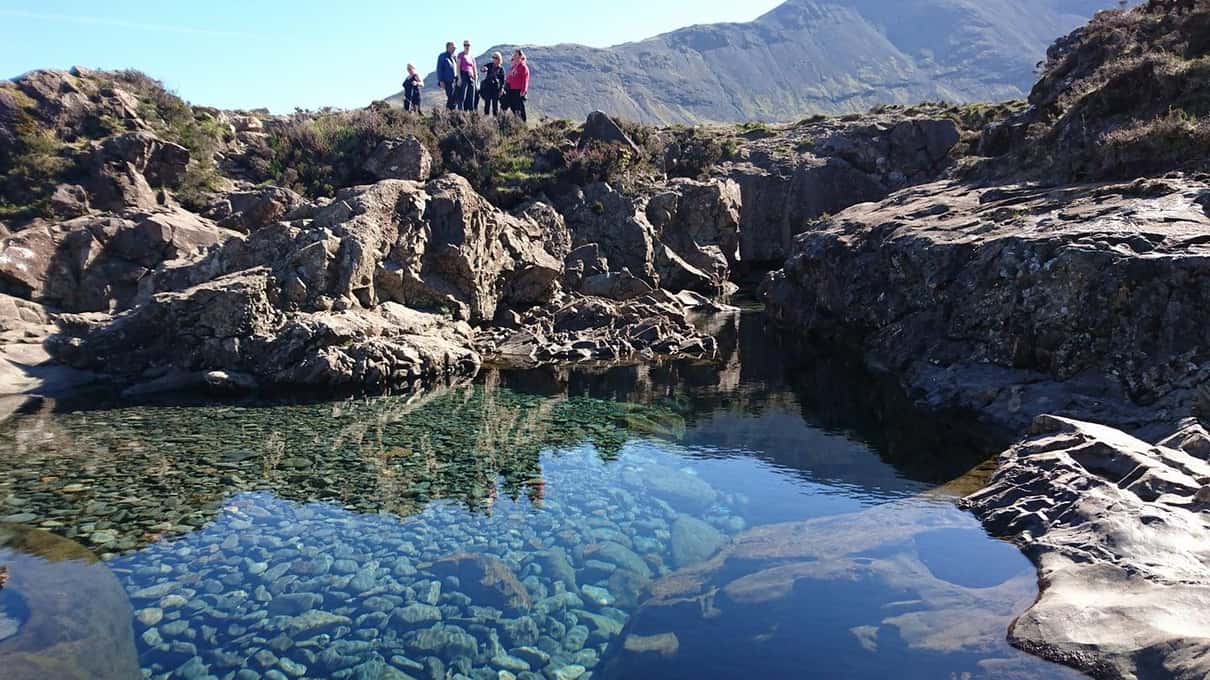 Walkers standing on the rocks above a clear turquoise Fairy Pool in Glen Brittle beneath the Black Cuillin