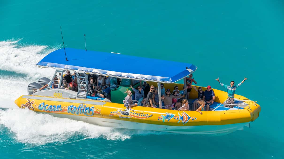A yellow Ocean Rafting inflatable with a blue canopy full of passengers speeding across blue water
