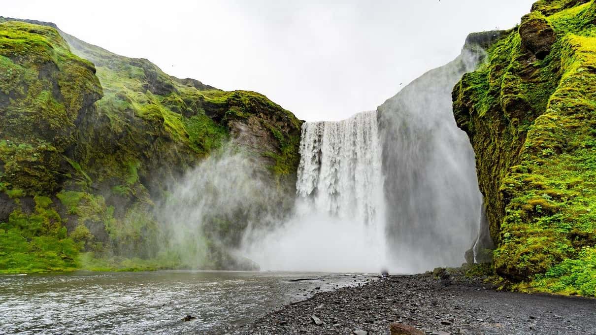 Der Wasserfall Skógafoss stürzt über eine moosgrüne Klippe in eine Wolke aus Gischt