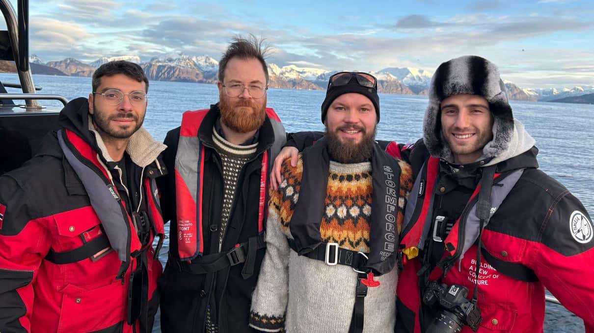 Four smiling guests in red flotation jackets and warm hats on a boat deck with a snowy coast behind them
