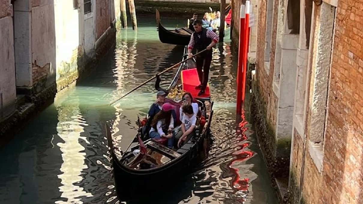 Gondel vol passagiers in een smal kanaal tussen bakstenen gevels, achterin roeit de gondelier langs rood-wit gestreepte meerpalen