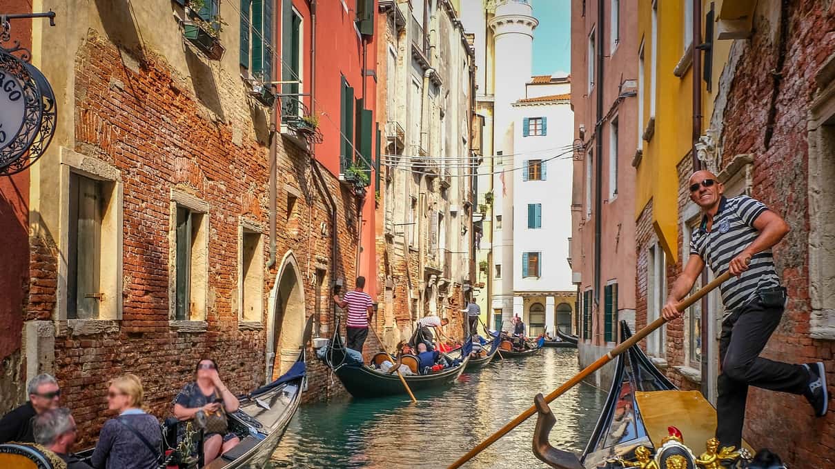 Gondolier en marinière qui prend appui du pied contre un mur dans un canal coloré, devant lui d’autres gondoles avec des passagers