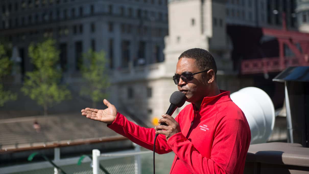 A docent in a red jacket pointing out buildings to passengers on the open upper deck of a river cruise boat