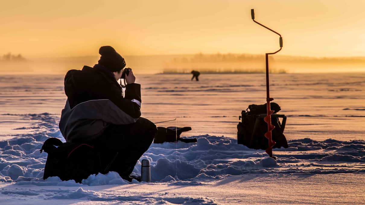 An angler crouched on a frozen lake near Levi at sunset beside an ice auger