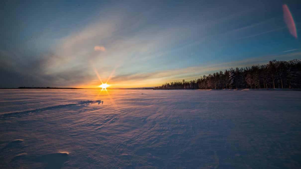 The sun setting over a wide frozen lake with a dark line of forest on the far shore