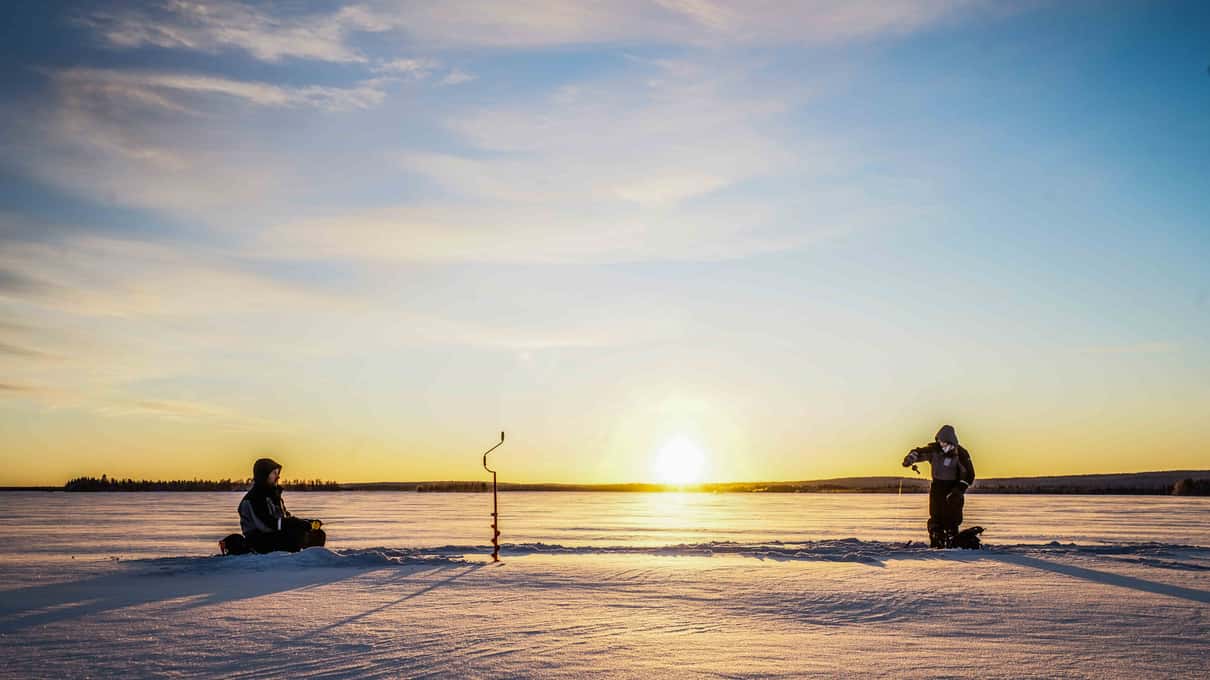 Two anglers sitting at their holes on a wide frozen lake under a low orange sun