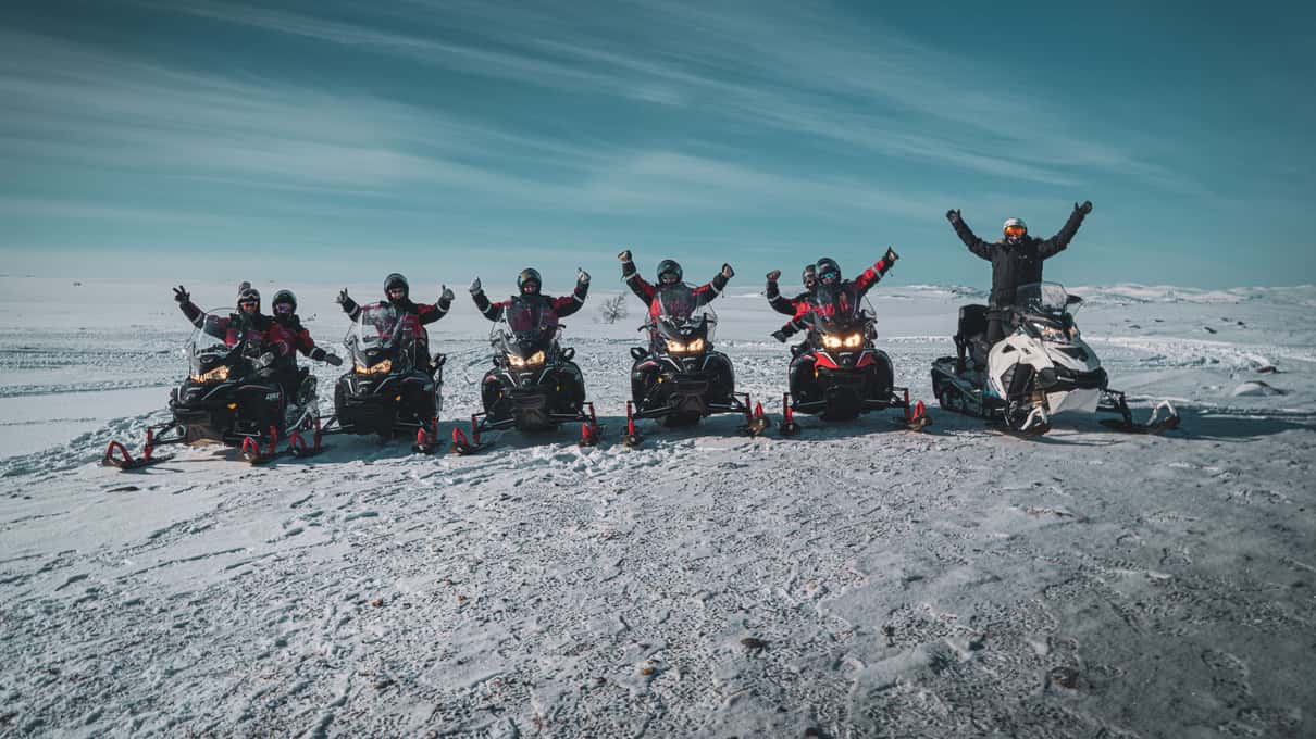 A row of riders in red suits on snowmobiles on the open plain above Gargia Valley