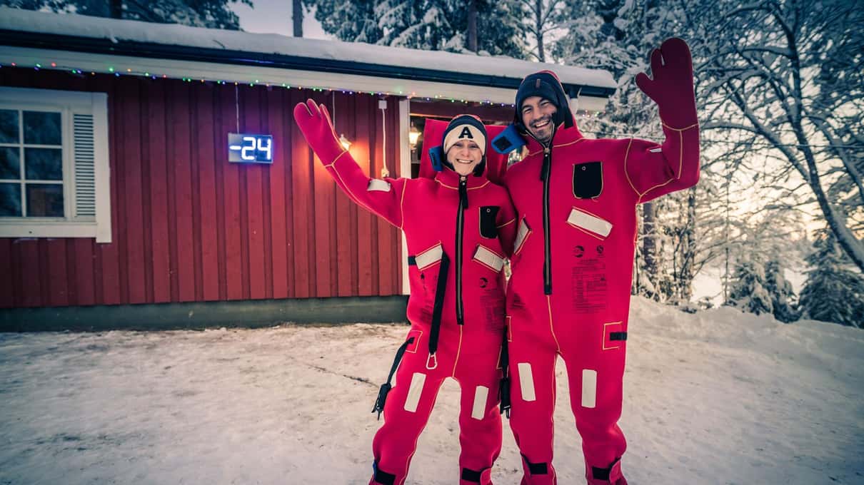 Two people in red rescue floating suits waving outside a red Lapland cottage in the snow