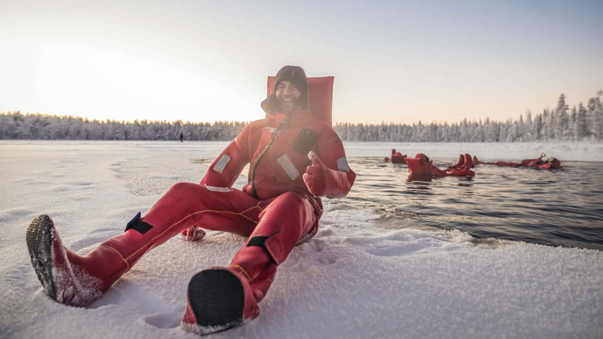 A floater in a red rescue suit sitting on the snowy edge of the ice hole while others float behind