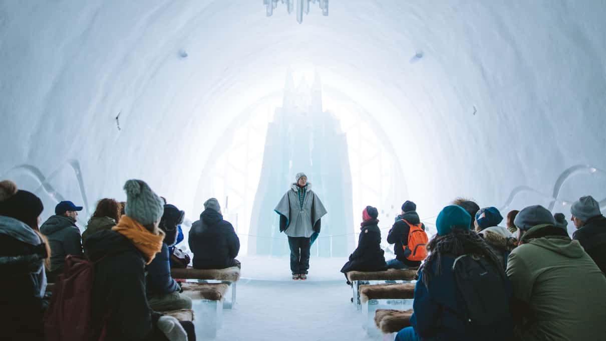 A guide speaking to visitors seated on ice benches in the vaulted Ice Church under an ice chandelier