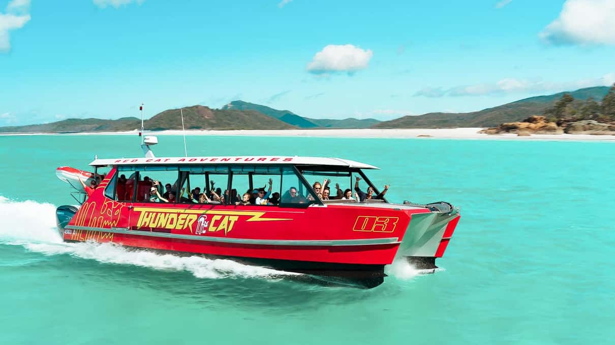 A red Red Cat Adventures catamaran full of passengers running at speed across turquoise water past a white sand beach
