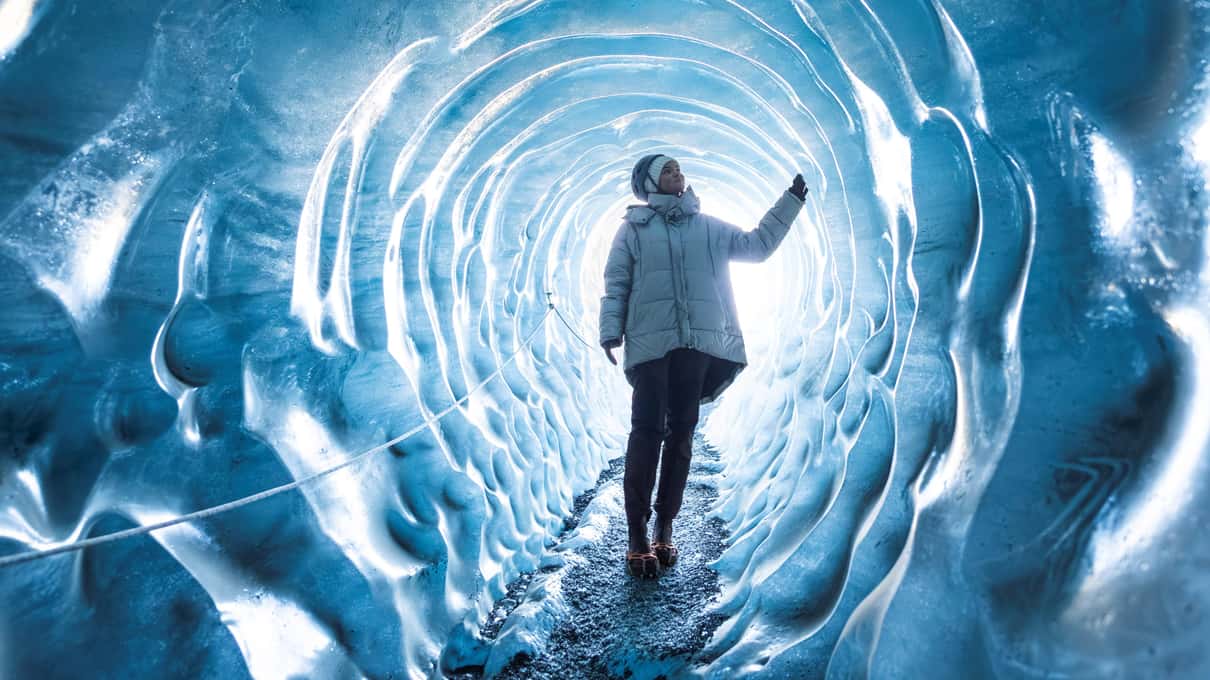 Un visiteur équipé de crampons marchant vers la lumière dans un tunnel de glace bleue ondulée