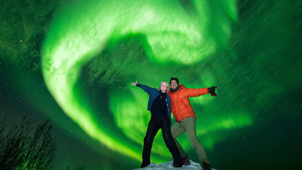 Deux voyageurs bras levés dans la neige sous un tourbillon d’aurore boréale verte