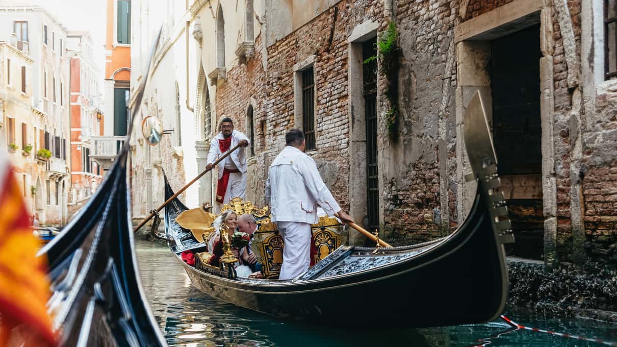 Deux gondoliers en tenue blanche rament une gondole noire décorée le long d’un mur de briques dans un canal étroit