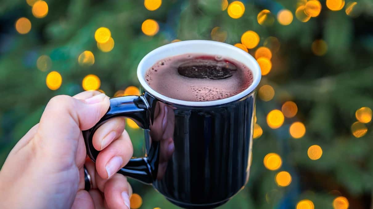 A hand holding a black mug of steaming mulled wine in front of a background of blurred fairy lights