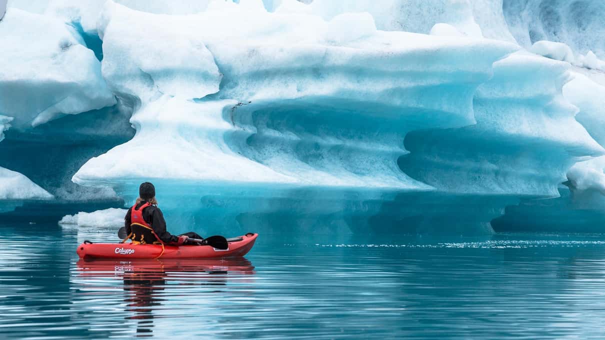 Kayaker in a red kayak paddling beneath a large blue iceberg on Jökulsárlón glacier lagoon
