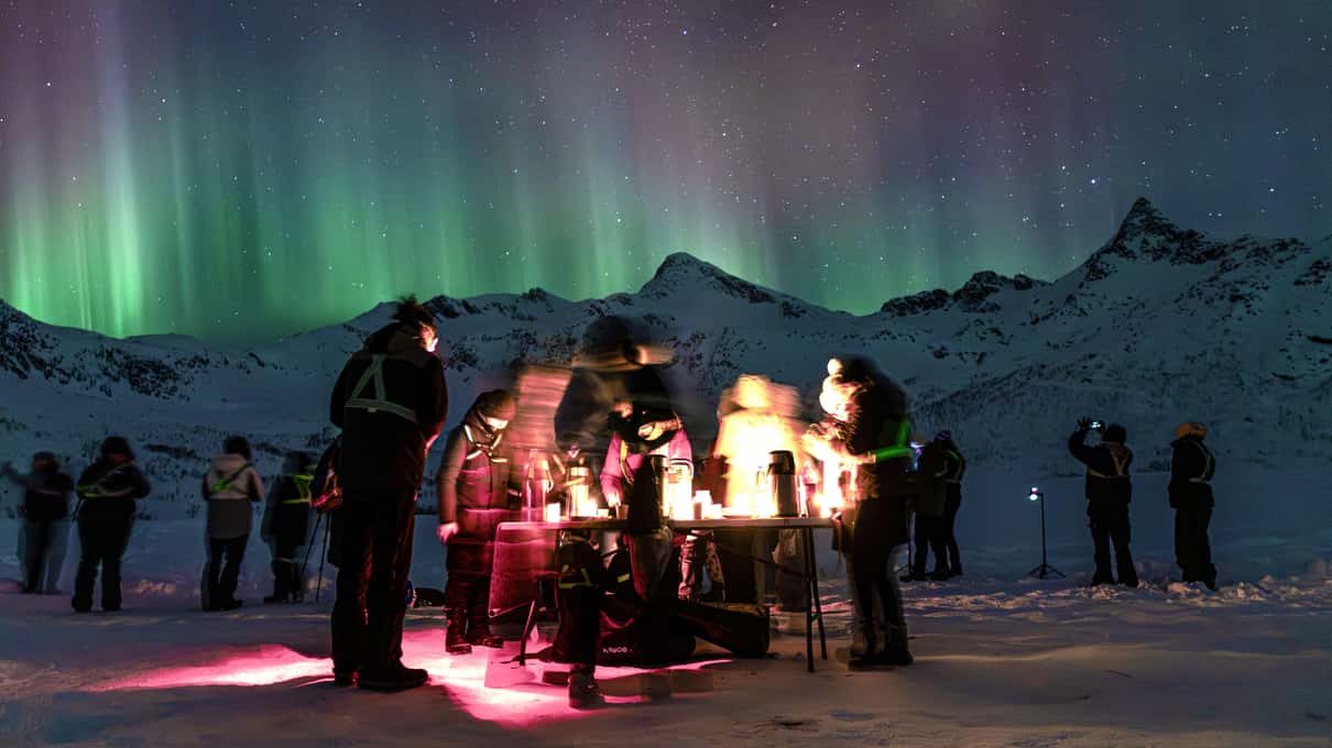 A group of guests around a campfire on snowy ground, green aurora curtains above the mountains